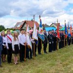 Grupa ludzi z flagami stoi na trawie, ubrani są odświętnie. W tle domy i namioty, niebo pokryte chmurami. Kolory flag są różnorodne.