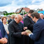 Grupa ludzi na zewnętrznej ceremonii, jasne niebo z chmurami. Kapłan w czarnej sutannie odbiera chleb. Tło z zielonymi namiotami i domami.
