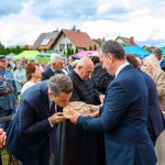 Na obrazie widać uroczystość plenerową z udziałem wielu osób. Mężczyzna całuje tradycyjny chleb, trzymany przez innego mężczyznę. W tle namioty i zachmurzone niebo.