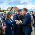 Na zdjęciu grupa ludzi w eleganckich ubraniach dzielących chleb. Tło to niebo z chmurami i domy z czerwonymi dachami. Wydarzenie odbywa się na świeżym powietrzu.