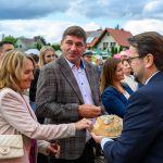 Grupa osób na zewnątrz uczestniczy w wydarzeniu. Kobieta w jasnym płaszczu łamie chleb. W tle domy i pochmurne niebo.