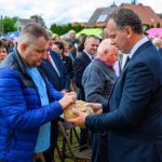 Mężczyźni ubrani w garnitur i niebieską kurtkę dzielą się chlebem na zewnątrz. Tło to tłum ludzi i kolorowe parasole. Atmosfera jest uroczysta.