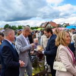 Grupa ludzi w eleganckich strojach uczestniczy w wydarzeniu plenerowym. Niebo jest pochmurne, a w tle widać namioty i domy. Osoba w garniturze wręcza kawałek chleba innemu uczestnikowi.