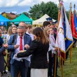 Grupa ludzi na uroczystości plenerowej, w tle kolorowe namioty i flagi. Osoby są elegancko ubrane. Tło to zielona trawa i drzewa.