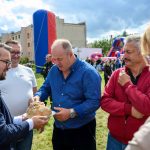 Grupa mężczyzn na trawie, jeden trzyma ozdobny bochenek chleba. W tle niebiesko-czerwona dmuchana atrakcja. Budynek pod chmurami.