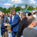 Na zdjęciu jest grupa osób na zewnątrz podczas wydarzenia. Mężczyzna w granatowym garniturze trzyma chleb. Tło jest zielone z flagami i niebieskim niebem.