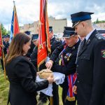 Grupa strażaków w mundurach stoi na trawie, trzymając sztandary. Kobieta podaje im chleb na białym obrusie. Tło to budynki i niebo z chmurami.