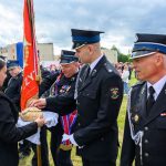 Uroczystość na świeżym powietrzu. Strażacy w mundurach przy chorągwiach, odbierają chleb od kobiety. Tłum w tle, pochmurne niebo.