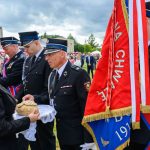 Ceremonia strażacka na świeżym powietrzu, mężczyźni w mundurach obok kobiety z chlebem. Czerwone sztandary dominują scenę. Tło to chmury i zielone drzewa.