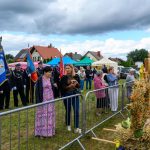 Na zdjęciu widać strażaków w mundurach z flagami oraz tłum ludzi. Tło to wiejskie domy i stoiska w kolorowych namiotach. Na pierwszym planie są dekoracje z kłosów zbóż i kwiatów.