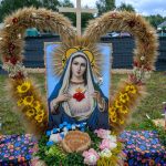 Obraz przedstawia wizerunek Maryi z otoczką z kłosów i słoneczników. Na dole kompozycja z kolorowych kwiatów i napisem. W tle zielone drzewa i namioty.