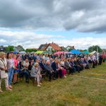Na zdjęciu widać tłum ludzi na zewnątrz w słoneczny dzień, siedzących i stojących przed sceną. W tle są kolorowe namioty, a niebo jest zachmurzone. Ludzie są ubrani w różnorodne stroje, a niektórzy trzymają flagi.