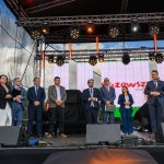 Grupa ludzi stoi na scenie pod czarnym zadaszeniem. Tło to wielki ekran z napisem w czerwieni. Prowadzący trzyma mikrofon.
