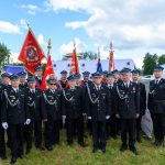 Grupa strażaków w czarnych mundurach z niebieskimi czapkami stoi na trawie. Za nimi widać kilka kolorowych flag. W tle błękitne niebo i drzewa.