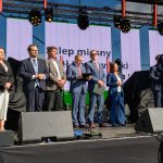 Na scenie stoi grupa osób w eleganckich strojach. W tle jasne ekrany z napisami. Na scenie sprzęt nagłośnieniowy.