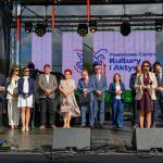 Na scenie stoi grupa osób w eleganckich strojach. Tło to duży ekran z napisem Powiatowe Centrum Kultury. Scena oświetlona jest jasnym światłem dziennym.