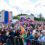 Tłum ludzi na zielonej łące przy kolorowych dmuchańcach. W tle bloki mieszkalne i drzewa. Niebieskie niebo z białymi chmurami.