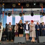 Na scenie stoi grupa ludzi w eleganckich ubraniach. Tło stanowi duży ekran z napisem i światła sceniczne. Niebo jest częściowo widoczne.
