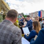 Na zdjęciu ludzie zgromadzeni wokół ozdoby ze zbóż. W tle budynki, niebo z chmurami, dominują kolory niebieski i zielony. Atmosfera wydarzenia plenerowego.