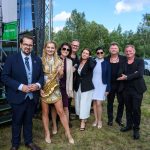 Grupa ludzi pozuje na zewnątrz na tle zielonych drzew. Kobieta w złotej sukience trzyma saksofon. Niebo jest błękitne z białymi chmurami.