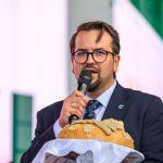 Mężczyzna w okularach trzyma chleb na scenie, mówi do mikrofonu. Ubrany w garnitur, w tle zielono-białe wzory. Na chlebie dekoracje z ciasta.