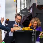 Na scenie troje ludzi w eleganckich ubraniach kroi chleb. Obok znajdują się słoneczniki na czarnym stole. W tle widać sprzęt nagłaśniający.
