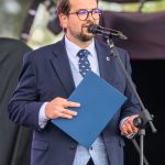 Mężczyzna w granatowym garniturze trzyma niebieski dokument i mówi do mikrofonu na scenie. Tło rozmyte, jasne kolory.