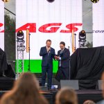 Dwóch mężczyzn w garniturach na scenie z mikrofonem i notatnikiem. Tło z jasnymi ekranami. Widownia przed sceną.
