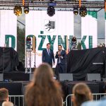 Na scenie, na tle dużego ekranu LED, dwaj mężczyźni w garniturach przemawiają. Ekran pokazuje napis Dożynki i symbol kłosa. Scena jest oświetlona reflektorami.