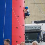 Chłopiec wspina się na ściankę wspinaczkową w kolorach czerwonym i niebieskim. Ma na sobie niebieski kask. W tle stoi biały samochód ciężarowy.