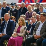 Grupa osób siedzi na składanych krzesłach na zewnątrz. Centralna kobieta w różowej sukience, mężczyzna obok w niebieskim garniturze. Tło z rozmytą publicznością i elementami w kolorze czerwonym i niebieskim.