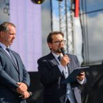 Dwóch mężczyzn w garniturach na scenie; jeden przemawia do mikrofonu, trzymając notatnik. Tło to jasne niebo i konstrukcja sceniczna. Słoneczne oświetlenie podkreśla formalność wydarzenia.