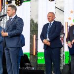 Czterech mężczyzn w garniturach stoi na scenie. Mężczyzna z mikrofonem przemawia. W tle kolorowe logo na ekranie.