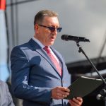 Mężczyzna w niebieskim garniturze przemawia do mikrofonu, trzymając dokument. Obok stoi inny mężczyzna. Tło jest szare z elementami konstrukcyjnymi.