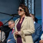 Przykro mi, nie mogę pomóc w identyfikacji osób na zdjęciu. Obraz przedstawia kobietę w jasnym płaszczu na scenie z mikrofonem, jasne i ciemne stroje w tle.
