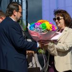 Kobieta w okularach przeciwsłonecznych przyjmuje bukiet kolorowych kwiatów od mężczyzny w garniturze. W tle mikrofony i ciemne niebo.