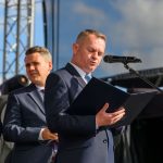 Trzech mężczyzn w garniturach, jeden z mikrofonem i dokumentem, tło z jasnym niebem i konstrukcją sceny.