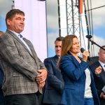 Grupa ludzi na scenie; kobieta w niebieskiej marynarce mówi do mikrofonu. Jasne niebo w tle, widoczna struktura nośna.