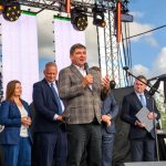 Na scenie stoi mężczyzna w marynarce, przemawiający do mikrofonu. W tle są jasne ekrany LED i konstrukcja sceniczna. Obok niego stoi grupa ludzi w garniturach.