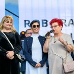Grupa osób na scenie, jedna kobieta mówi do mikrofonu. Zróżnicowane kolory ubrań, tło z napisem. Atmosfera formalna.