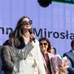 Na scenie stoi kobieta w białym płaszczu z mikrofonem, uśmiecha się. W tle duży ekran z tekstem i niebo. Obok niej kilka osób w formalnych strojach.
