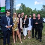 Grupa ludzi na trawie w eleganckich strojach, kobieta trzyma saksofon. W tle sceniczne rusztowanie i drzewa, niebo z chmurami.