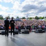 Na scenie w plenerze dwóch mężczyzn i kobieta z saksofonem patrzą na tłum. Jasne, słoneczne tło, niebo z chmurami. Za nimi widoczne namioty i kolorowe atrakcje.