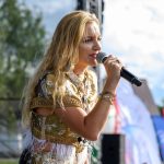 Kobieta na scenie śpiewa do mikrofonu, ubrana w złotą bluzkę. Scena jest na zewnątrz, w tle drzewa i niebo. Kolorowe flagi widoczne po prawej.
