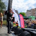 Koncert na scenie plenerowej, mężczyzna w garniturze z mikrofonem, kobieta w sukience, tłum ludzi i budynki w tle.