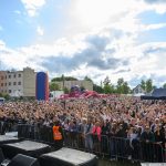 Tłum na koncercie na zewnątrz, ludzie wznoszą ręce, niebo jest częściowo pochmurne. Kolory: niebieski, czerwony, zielony. W tle budynki.