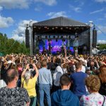 Tłum fanów przed sceną na koncercie plenerowym. Nad sceną czarne zadaszenie, tło z niebieskim niebem i chmurami. Ludzie unoszą ręce, scena oświetlona.