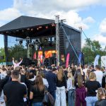 Koncert plenerowy z czarną sceną, kolorowymi światłami i tłumem widzów na tle błękitnego nieba oraz zieleni drzew.