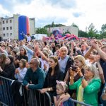 Tłum ludzi na plenerowym koncercie przy jasnym niebie. Kolorowe dmuchańce w tle przy blokach mieszkalnych. Ludzie entuzjastycznie klaszczą.