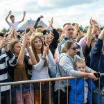 Grupa ludzi na koncercie, wznoszących ręce w radosnym geście. W tle widać niebo pełne chmur. Barwne ubrania tworzą żywą atmosferę.